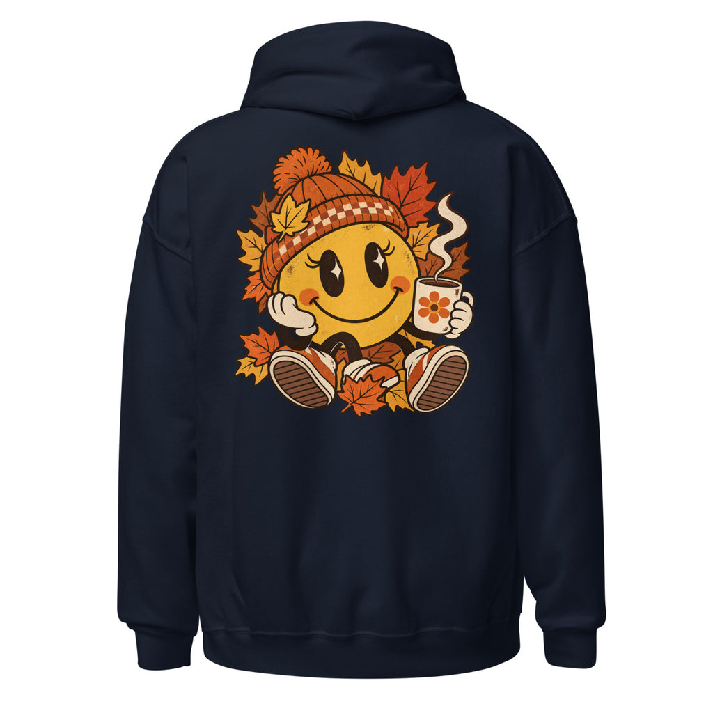 Lächelnder gelber Smiley mit großen glänzenden Augen, orange karierter Strickmütze und weißen Handschuhen sitzt umgeben von orange und gelb gefärbten Ahornblättern, hält eine dampfende Kaffeetasse mit Blumenmotiv in der rechten Hand. – Unisex Heavy Blend Hoodie | Gildan 18500