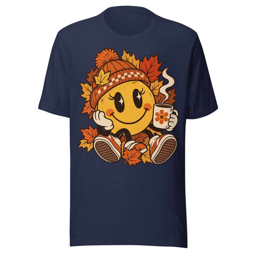 Lächelnder gelber Smiley mit großen glänzenden Augen, orange karierter Strickmütze und weißen Handschuhen sitzt umgeben von orange und gelb gefärbten Ahornblättern, hält eine dampfende Kaffeetasse mit Blumenmotiv in der rechten Hand. – Unisex Staple T-Shirt | Bella + Canvas 3001
